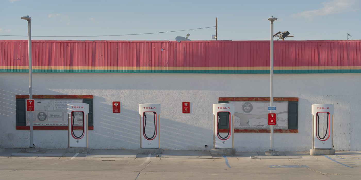 Tesla Supercharger V3 stalls in a row at a highway service station, posts lined up at dusk